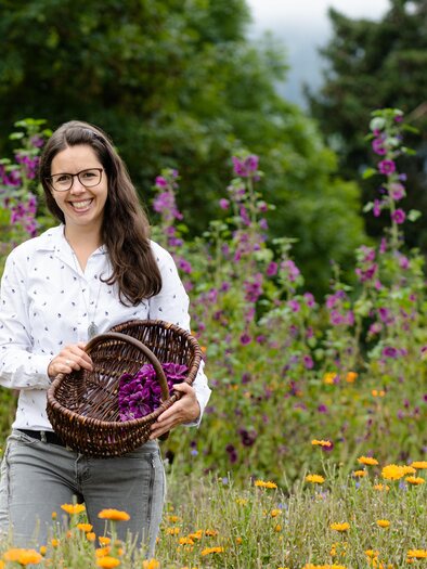 Eine Frau steht in einem bunten Blumenfeld und hält zwei Körbe mit Blumen. Im Hintergrund sind grüne Bäume und Nebel zu sehen. | © Martina Siebenhandl