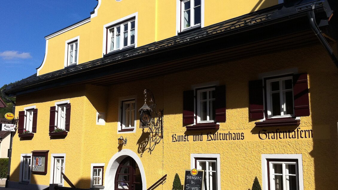 Ein gelbes Gebäude mit weißen Fenstern und roten Fensterläden. Es steht an einer Straße und hat ein einladendes Ambiente. | © Kunst- und Kulturhaus Öblarn