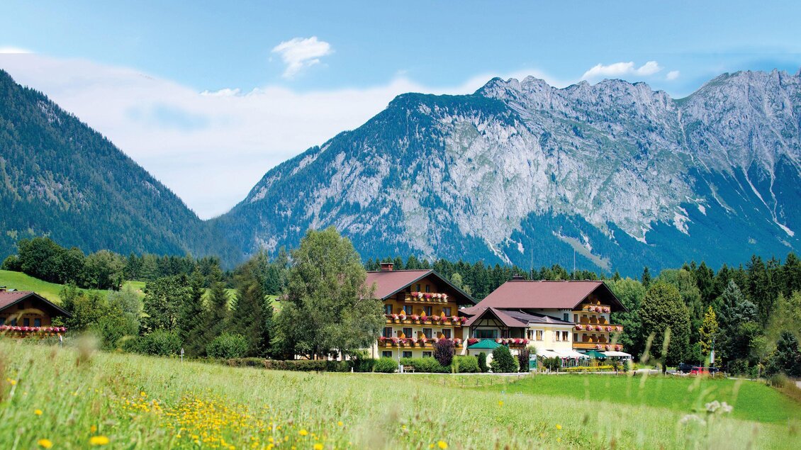 Ein malerisches Bergdorf mit traditionellen Häusern und einer blühenden Wiese. Im Hintergrund erheben sich beeindruckende Berge unter einem klaren Himmel. | © Landhotel - Restaurant Häuserl im Wald