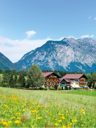 A picturesque mountain village with traditional houses and a blooming meadow. In the background, impressive mountains rise under a clear sky. | © Landhotel - Restaurant Häuserl im Wald