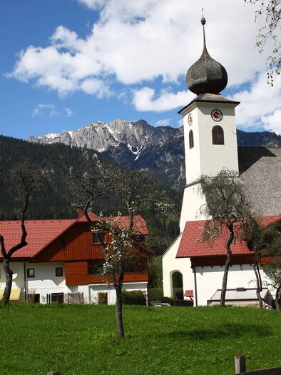 An idyllic landscape with a church and traditional buildings. In the background, the majestic mountains and a clear sky are visible. | © Ewald Gabardi