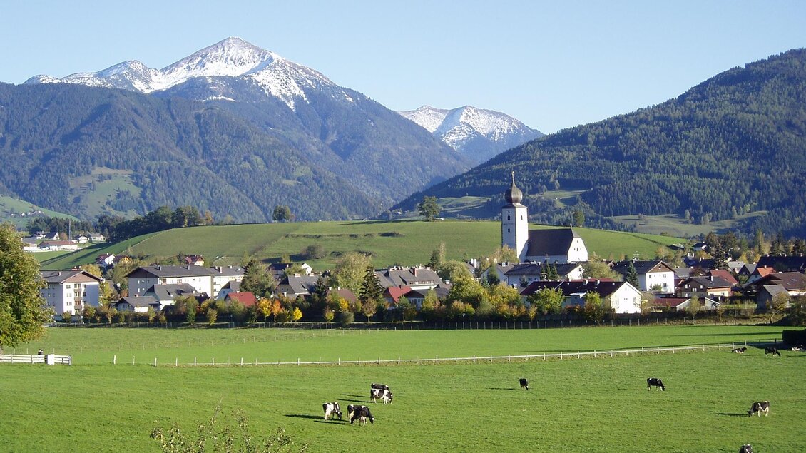 Eine malerische Landschaft mit grünen Wiesen und Kühen. Im Hintergrund sind schneebedeckte Berge und ein charmantes Dorf mit einer Kirche zu sehen. | © Marktgemeinde Gröbming