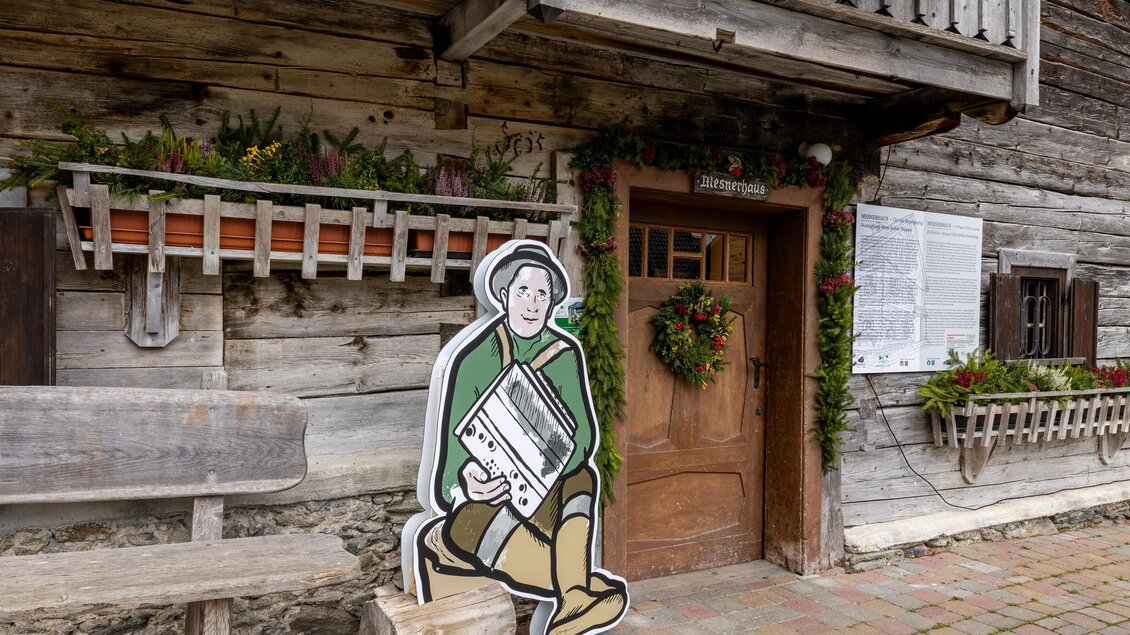 Ein traditionelles Holzhaus mit einer freundlichen Dekoration. Vor dem Eingang steht eine farbenfrohe Figur, die ein Musikinstrument spielt. | © Harald Steiner