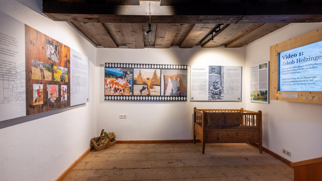 Ein Ausstellungsraum mit verschiedenen Kunstwerken und Informationsplakaten an den Wänden. In der Mitte steht ein historisches Möbelstück und ein Bildschirm zeigt ein Video. | © Harald Steiner