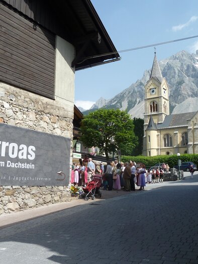 A picturesque street in Ramsau am Dachstein with a historic church in the background. People are gathered in front of a large sign that reads “Zeitroas.” | © Museum Zeitroas