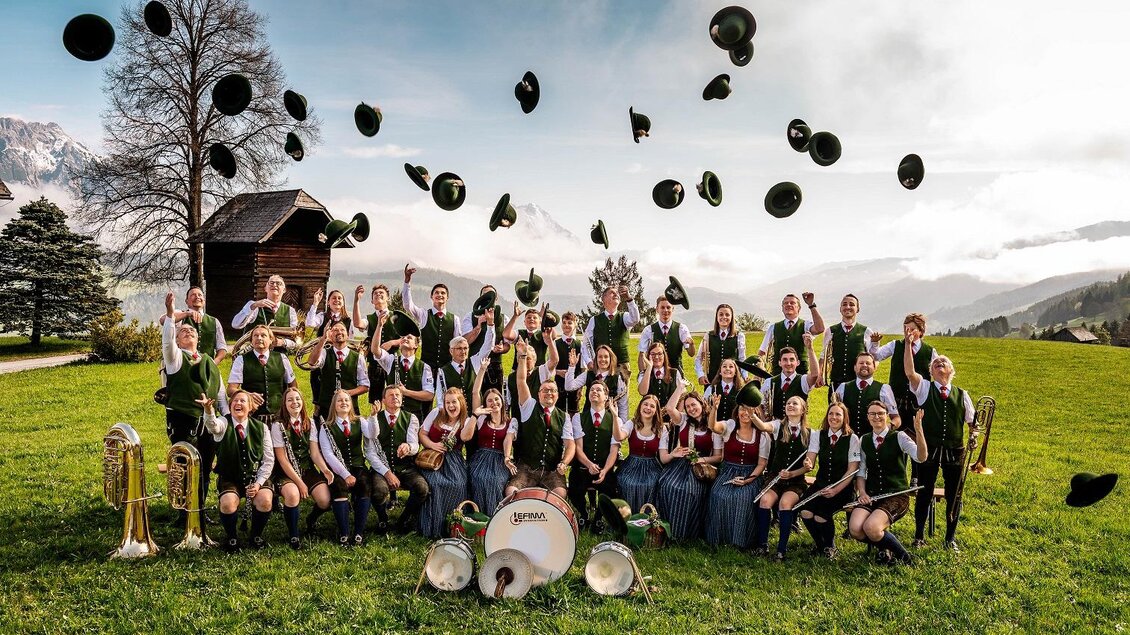 Eine große Gruppe von Musikern steht auf einer Wiese in den Bergen. Sie feiern mit ihren Hüten, die in die Luft geworfen werden. | © David Stocker