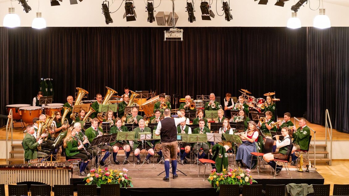 Eine Blaskapelle probt auf einer Bühne in einem Saal. Die Musiker tragen traditionelle Trachten und spielen verschiedene Instrumente. | © David Stocker