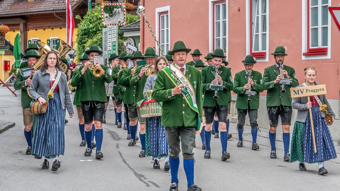 Eine traditionelle Blaskapelle marschiert durch eine malerische Straße. Die Musiker tragen Trachten und spielen verschiedene Instrumente. | © fotobook