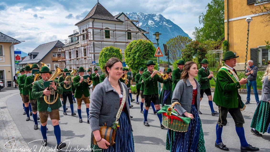 Eine traditionelle Parade mit Musikern in Tracht und Frauen in festlichen Kleidern. Im Hintergrund sind Berge und historische Gebäude zu sehen. | © fotobook