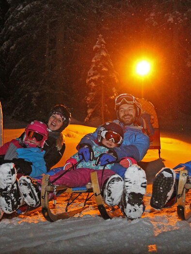 Eine Gruppe von vier Personen sitzt auf Schlitten im Schnee. Es ist dunkel, und ein Lichtschein beleuchtet die Szene. | © Hans-Peter Steiner