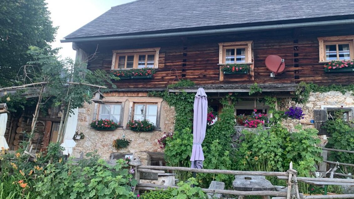 Ein charmantes Holzhaus mit blühenden Pflanzen und Fenstern mit Blumenkästen. Das Gebäude hat eine einladende Atmosphäre und liegt umgeben von üppigem Grün. | © Oberkogler-Alm