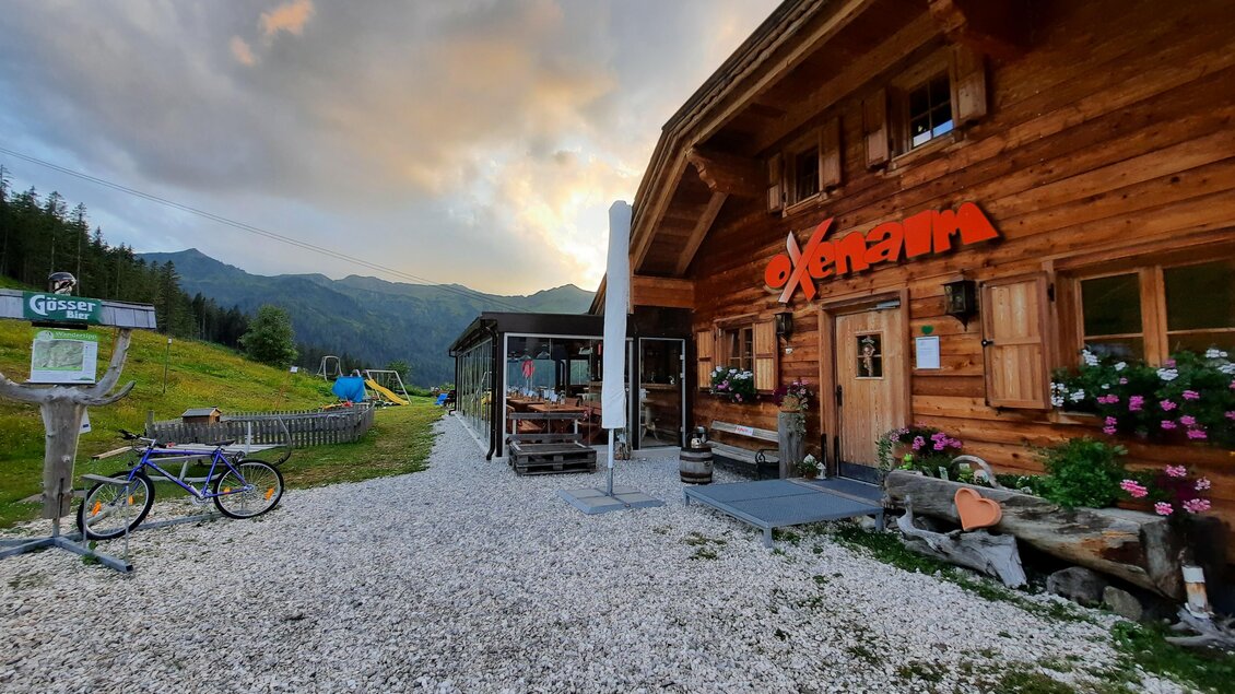 Ein rustikales Holzgebäude mit dem Namen "Kenzmann" steht in einer malerischen Berglandschaft. Umgeben von grünen Wiesen und Bergen, lädt es zum Verweilen ein. | © Oxenalm