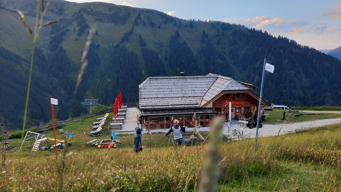 Eine charmante Almhütte umgeben von Bergen und einer schönen Wiese. Im Vordergrund sind Menschen zu sehen, die die Aussicht genießen. | © Oxenalm