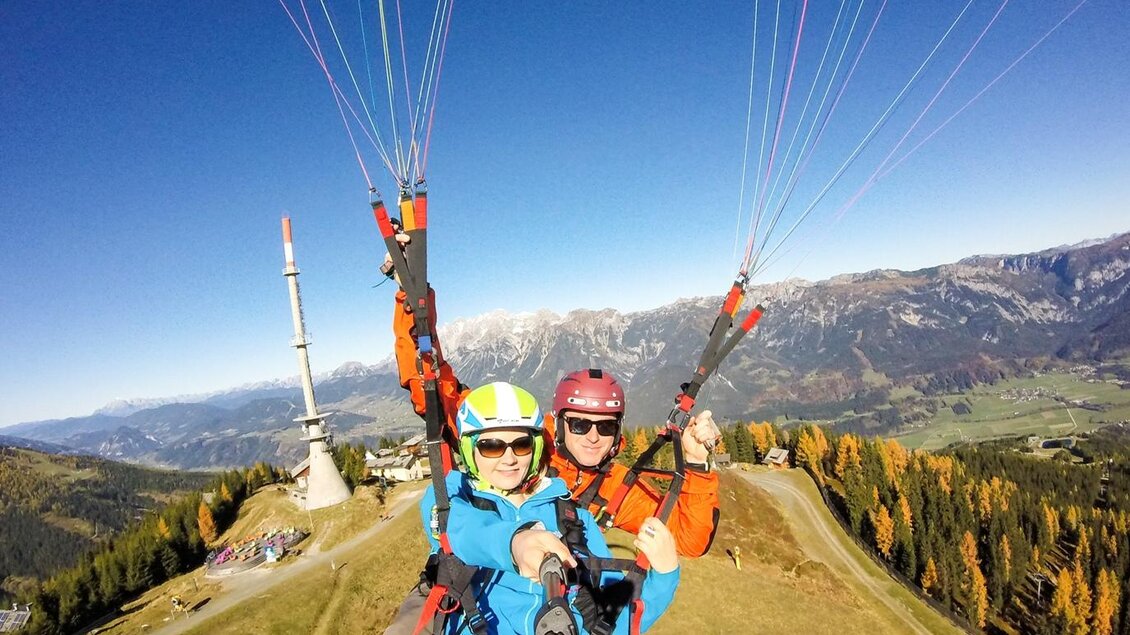Zwei Personen beim Paragliding, mit atemberaubendem Blick auf die Berge und die Landschaft. Die Umgebung ist grün und die Berggipfel sind im Hintergrund zu sehen. | © Flugschule Aufwind 