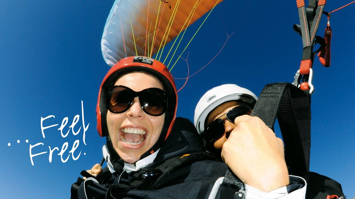 Ein Paraglider mit zwei Personen, die glücklich in die Kamera lächeln. Im Hintergrund ist ein klarer blauer Himmel zu sehen. | © Firma Nova 