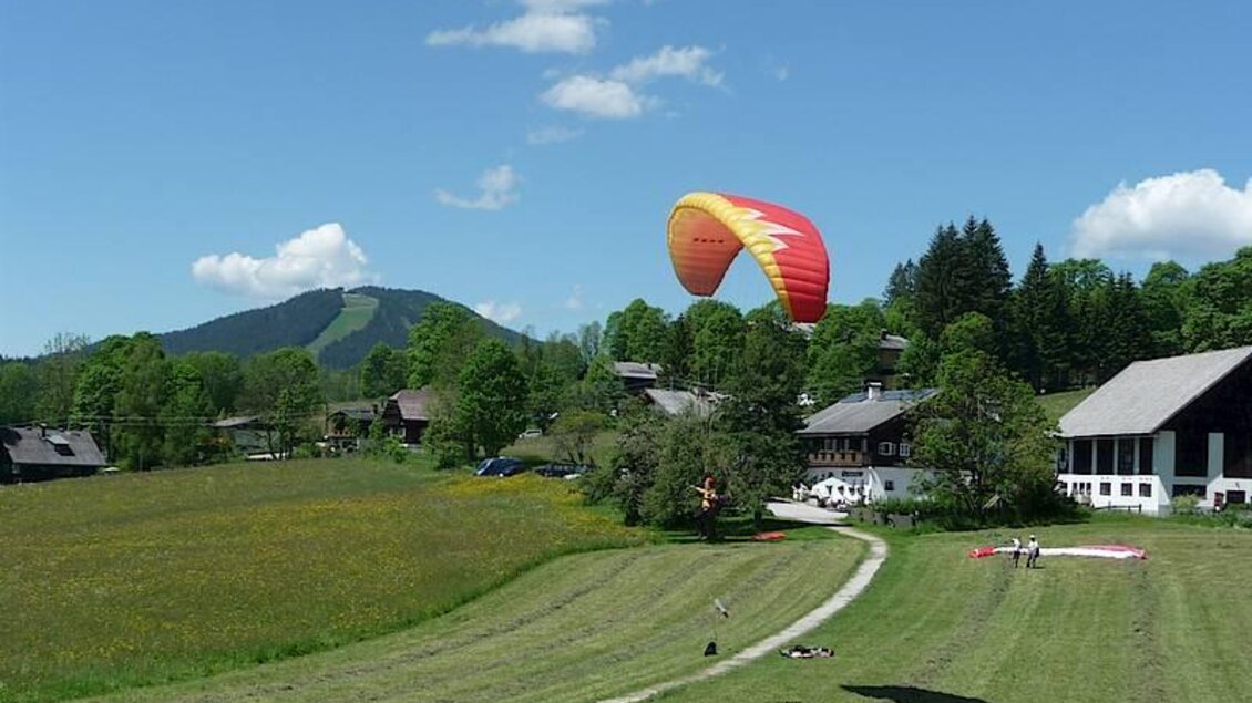 Ein Paraglider schwebt über eine grüne Wiese mit verstreuten Häusern und sanften Hügeln im Hintergrund. Der Himmel ist klar und blau mit wenigen Wolken. | © Flugschule Aufwind 