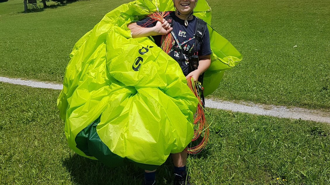 Eine Person steht auf einer Wiese und hält einen großen, leuchtend grünen Paraglider. Im Hintergrund sind Bäume und ein klarer Himmel zu sehen. | © Flugschule Aufwind 