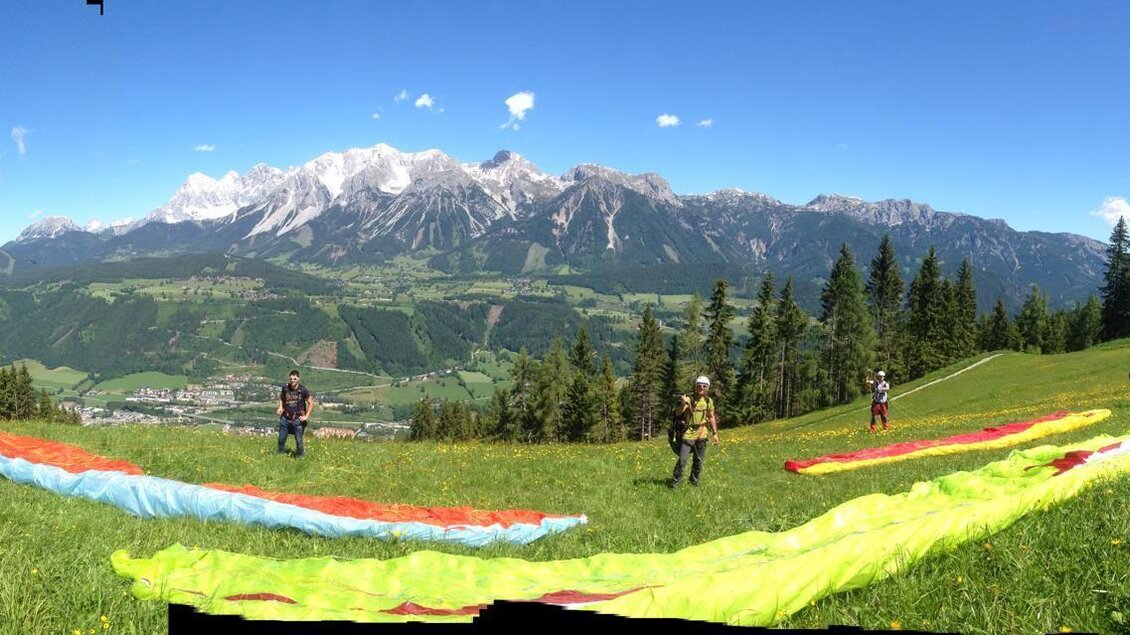 Eine malerische Landschaft mit grünen Wiesen und hohen Bergen im Hintergrund. Mehrere Personen bereiten sich darauf vor, mit bunten Gleitschirmen zu starten. | © Flugschule Aufwind 