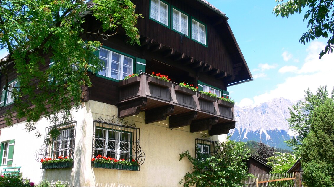 Ein traditionelles, mehrstöckiges Haus mit einem Balkon und bunten Blumen. Im Hintergrund sind die Berge und ein klarer Himmel zu sehen. | © Gernot Langs