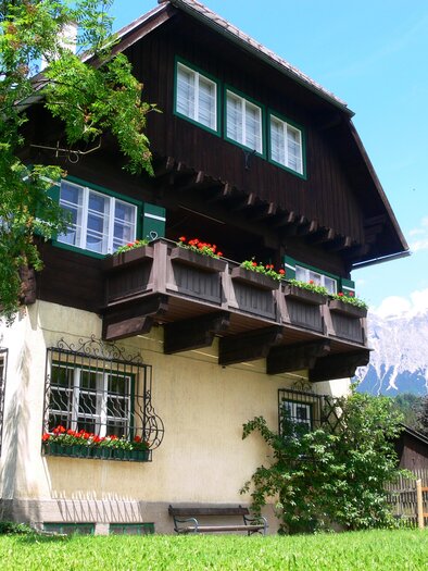 A traditional multi-story house with a balcony and colorful flowers. In the background, the mountains and a clear sky can be seen. | © Gernot Langs