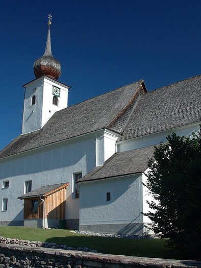 A beautiful church with a tall, pointed tower and traditional architecture. In the background, there are some houses and trees, and the sky is clear and blue. | © TVB Haus-Aich-Gössenberg