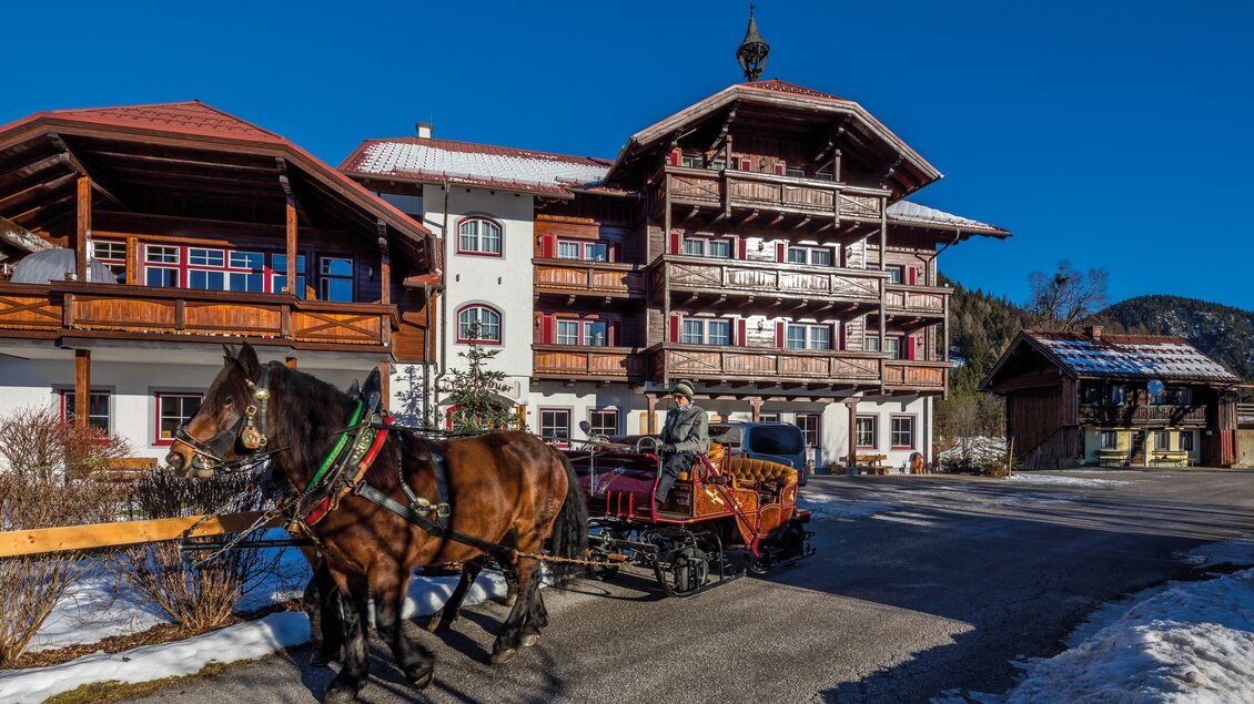 Eine Pferdekutsche fährt vor einem traditionellen Gebäude mit Holzverzierungen. Der Himmel ist klar und blau, und die Umgebung ist schneebedeckt. | © Peterbauerhof