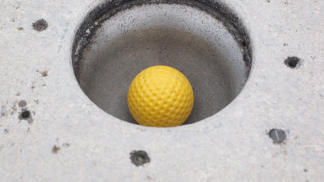 Eine gelbe Golfball liegt in einem Betonloch. Der Hintergrund ist grau und schlicht. | © Puttererseehof