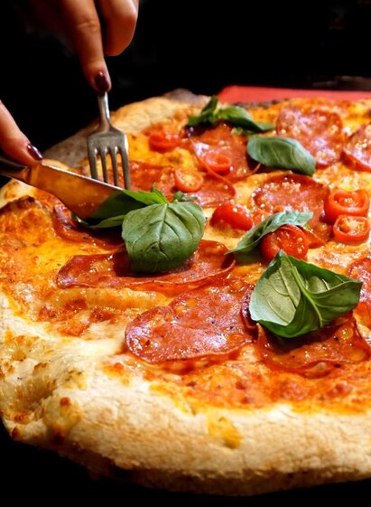 Eine frisch zubereitete Pizza mit Salami, Kirschtomaten und frischem Basilikum. Eine Person schneidet die Pizza mit Messer und Gabel. | © Symbolfoto
