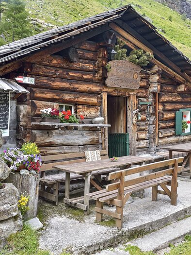 Eine rustikale Berghütte aus Holz mit Blumenschmuck. Draußen stehen Holztische und -bänke, umgeben von einer schönen Landschaft. | © Herfried Marek
