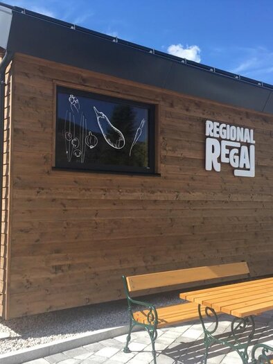Ein modernes Holzgebäude mit der Aufschrift "REGIONAL REGAL". Vor dem Gebäude stehen Tische aus Holz. | © Regional Regal