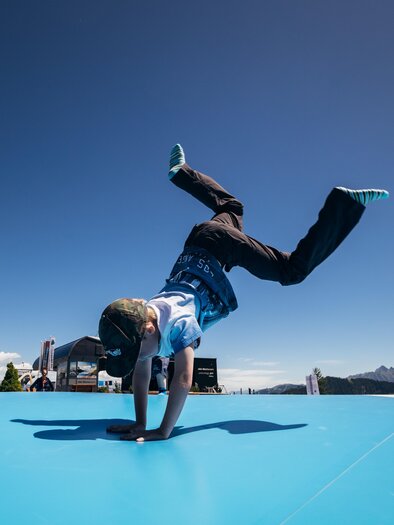 A boy is doing a handstand on a blue surface. In the background, there are mountains and a clear sky visible.