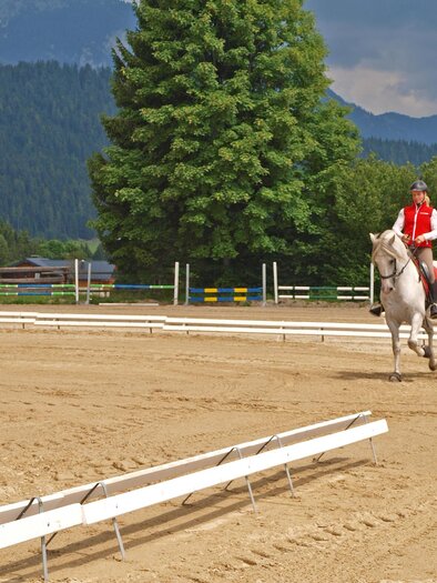 A rider in a red vest is riding a white horse in a sand arena. In the background, trees and mountains can be seen. | © Reiterhof Brandstätter