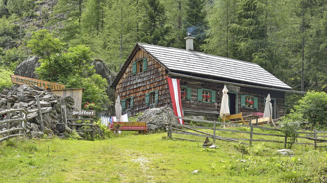 Eine rustikale Hütte mit Holzfassade inmitten von grünen Bäumen. Tische und Stühle stehen draußen, umgeben von einer ländlichen Atmosphäre. | © Herfried Marek
