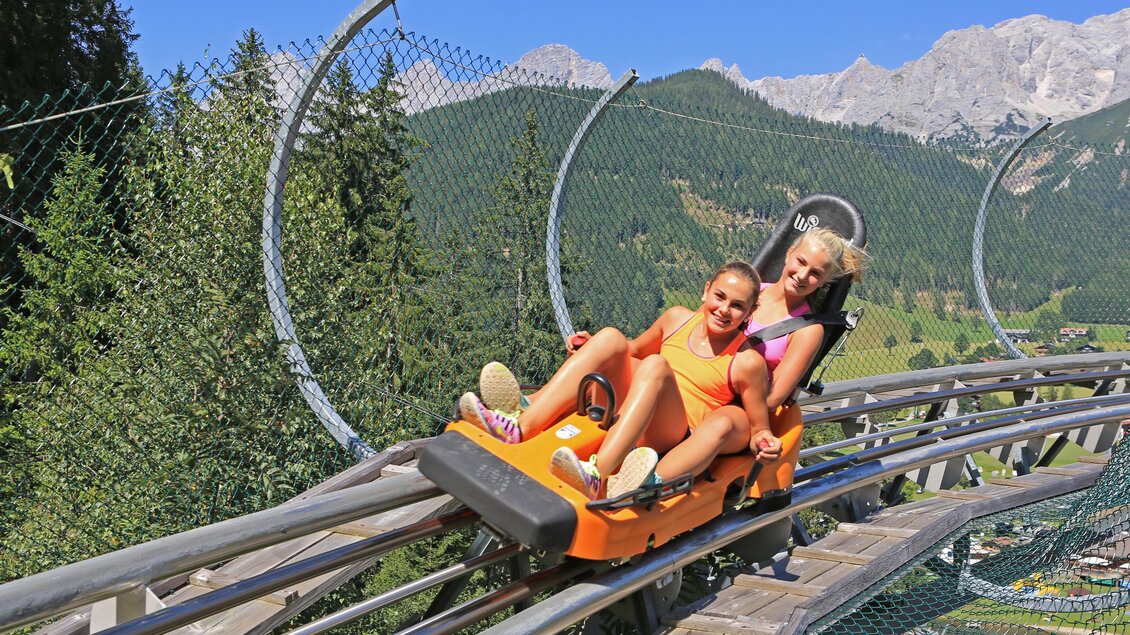 Zwei Frauen fahren fröhlich auf einer Sommerrodelbahn in den Bergen. Die Umgebung ist grün und die Berge sind im Hintergrund sichtbar. | © Erlebnis Rittisberg