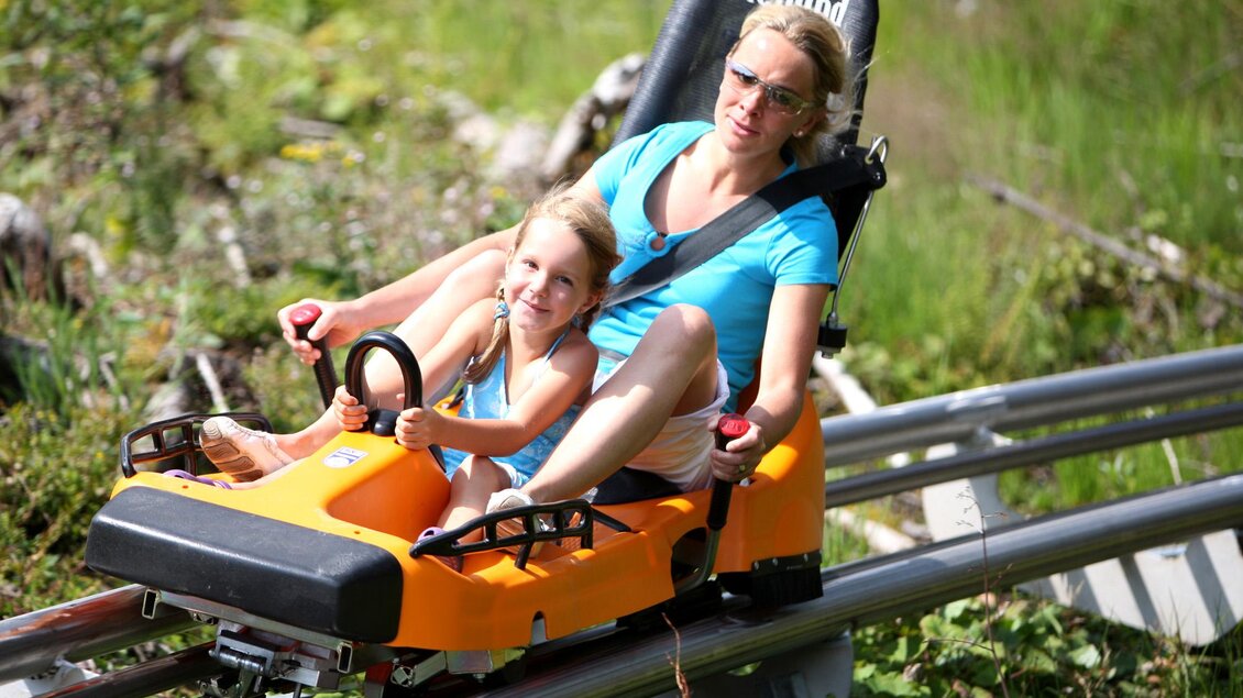 Eine Frau und ein Kind fahren gemeinsam auf einer Sommerrodelbahn. Die grüne Umgebung und die Sonne sorgen für eine fröhliche Stimmung. | © Erlebnis Rittisberg
