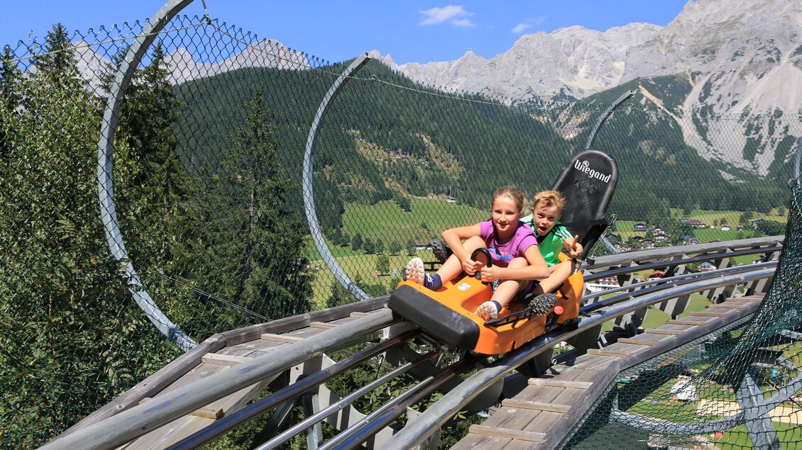 Zwei Kinder fahren auf einer Sommerrodelbahn in den Bergen. Im Hintergrund sind grüne Wälder und hohe Berge zu sehen. | © Erlebnis Rittisberg