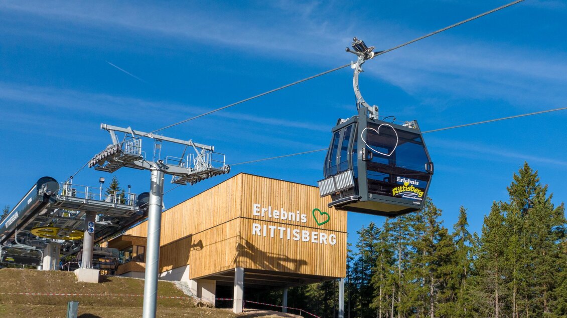 Eine Seilbahn fährt über eine grüne Wiese. Im Hintergrund befindet sich das Gebäude der Talstation mit Bäumen und blauem Himmel. | © Hans-Peter Steiner