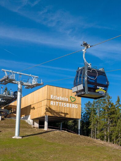 Eine Seilbahn fährt über eine grüne Wiese. Im Hintergrund befindet sich das Gebäude der Talstation mit Bäumen und blauem Himmel. | © Hans-Peter Steiner