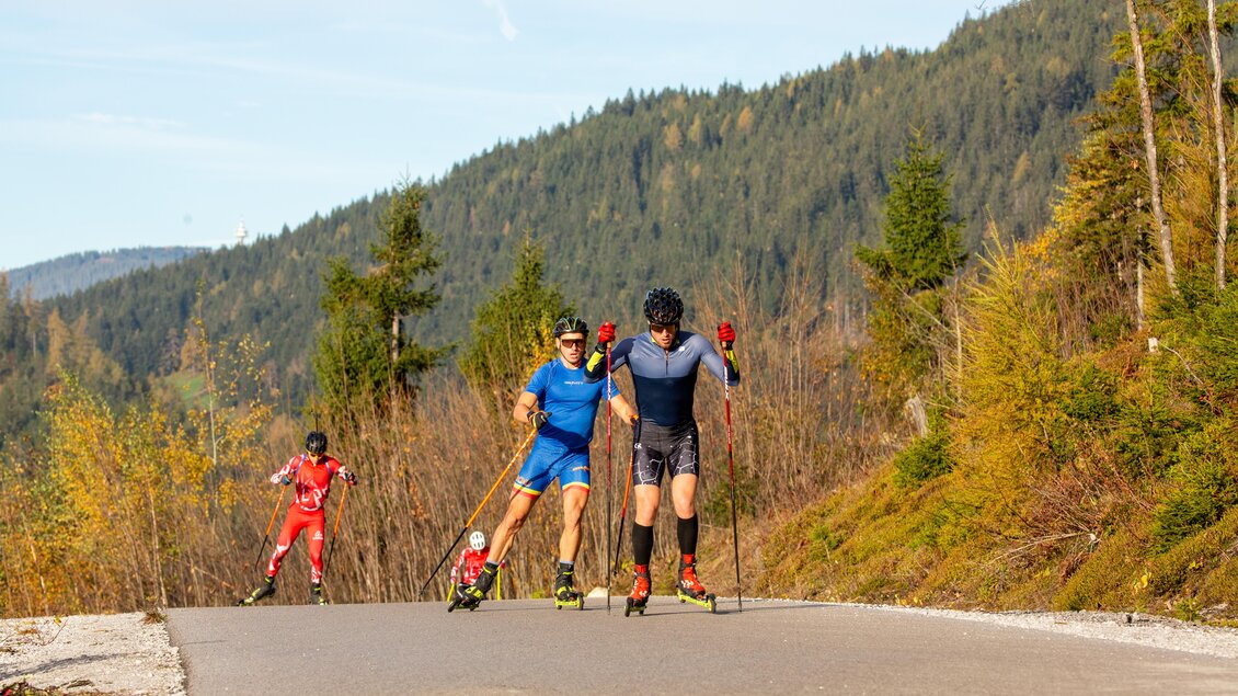 Zwei Männer in Sportkleidung trainieren mit Rollski auf einer Straße in den Bergen. Im Hintergrund ist ein dritter Sportler sichtbar.