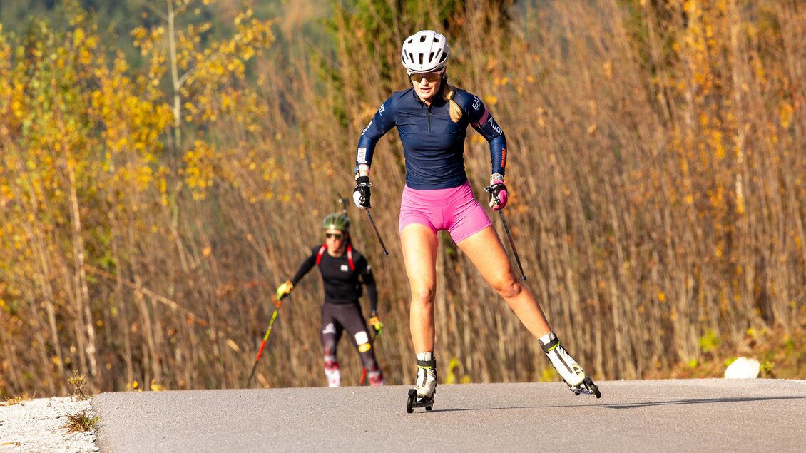 Zwei Sportlerinnen beim Rollerski im Herbst. Die Bäume im Hintergrund sind bunt gefärbt.