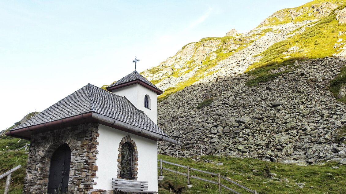 Eine kleine Kapelle steht am Fuß eines bewaldeten Berghangs. Im Hintergrund sind steile Felsen und grünes Gras sichtbar. | © Naturpark Sölktäler