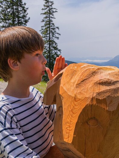 Zwei Kinder spielen mit Holzskulpturen in der Natur. Eine der Skulpturen stellt einen Hund dar und die Umgebung umfasst Bäume und Berge. | © Hauser Kaibling René Eduard Perhab