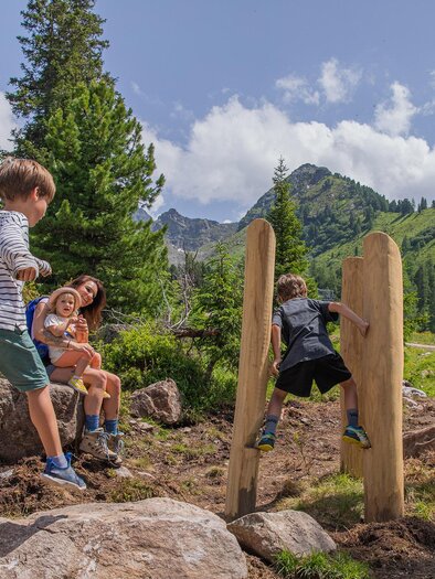 Eine Gruppe von Kindern spielt in der Natur auf einem Spielplatz aus Holz. Im Hintergrund sind grüne Bäume und Berge unter einem sonnigen Himmel zu sehen. | © Hauser Kaibling René Eduard Perhab