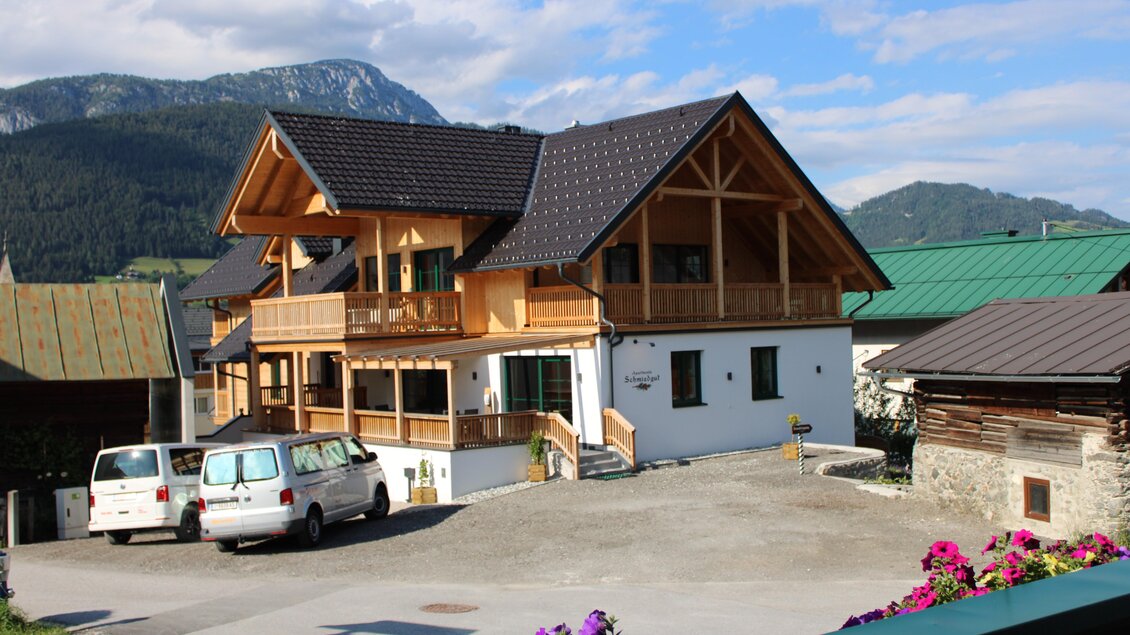 Ein schönes Holzhaus mit einem großen Balkon in einer bergigen Umgebung. Im Vordergrund sind parkende Autos und bunte Blumen zu sehen. | © Schmiedgut