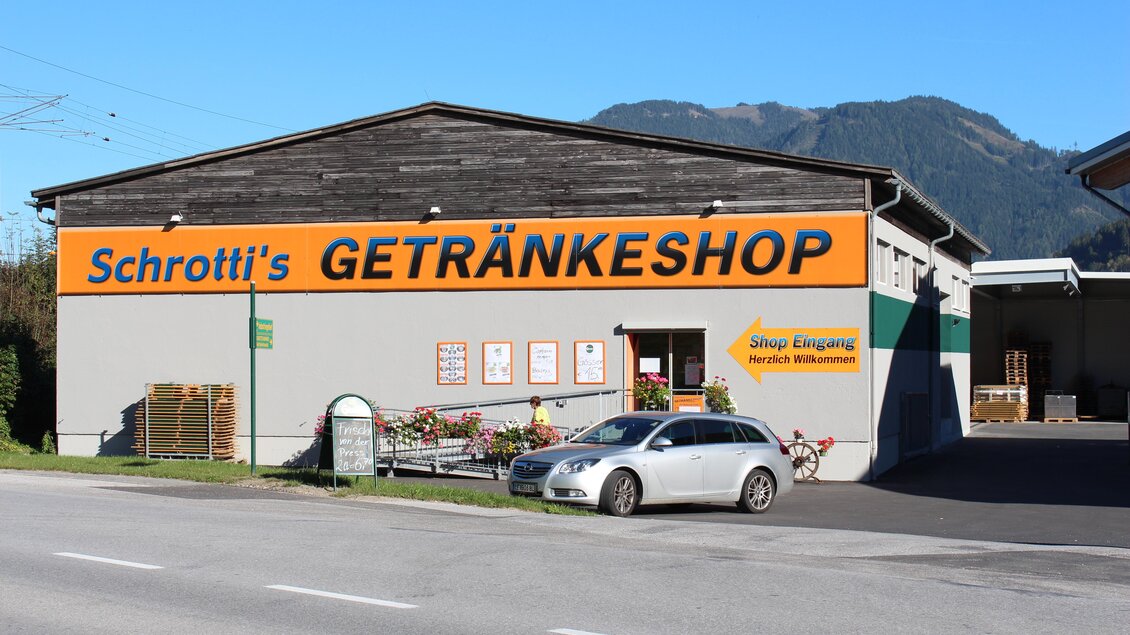 Ein Getränkeshop mit der Aufschrift "Schrotti's GETRÄNKESHOP" ist zu sehen. Vor dem Geschäft parkt ein Auto, und im Hintergrund sind Berge sichtbar. | © Schrotti's Getränkeshop