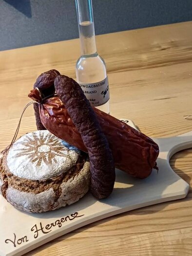 A rustic wooden board with farmhouse bread, sausage, and a small bottle. The arrangement conveys a hearty, traditional dining experience. | © Familie Gruber-Greil