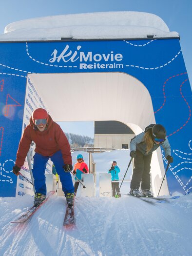 A group of skiers is skiing through a colorful arch in the ski resort. In the background, snow-covered trees and a clear sky can be seen.