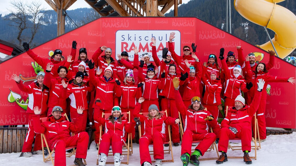 Eine fröhliche Gruppe von Skilehrern in roten Anzügen posiert vor einem großen Schild der Skischule Ramsau. Sie stehen auf Schnee und zeigen ihre Begeisterung mit gehobenen Händen. | © Skischule | Alpincenter Dachstein