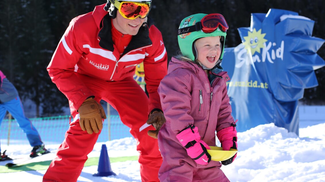 Ein Skilehrer hilft einem fröhlichen kleinen Mädchen beim Skifahren. Beide tragen Schutzkleidung und stehen auf Schnee, umgeben von anderen Skifahrern. | © Kinderskikurs | Alpincenter Dachstein
