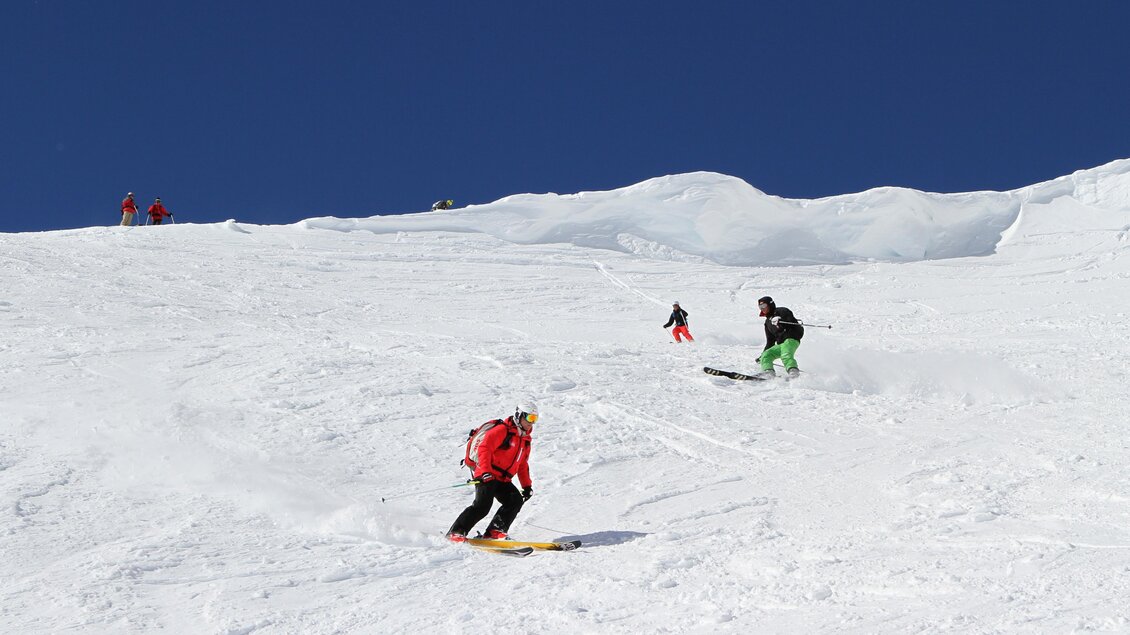 Zwei Skifahrer fahren auf einer schneebedeckten Piste. Im Hintergrund sind weitere Personen und ein klarer blauer Himmel zu sehen. | © Abfahrt auf der Piste | Alpincenter Dachstein